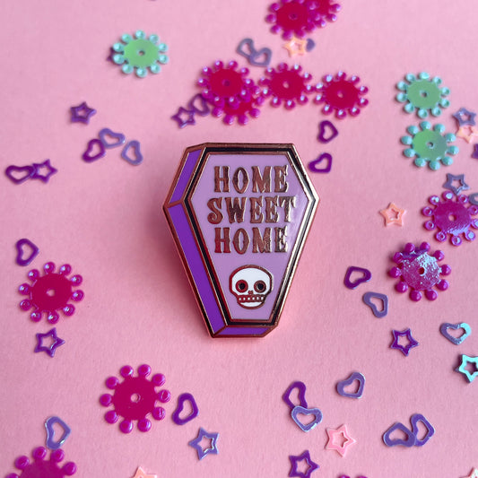 A coffin shaped enamel pin with the words "Home Sweet Home" on it and a skull on a pink background with confetti around it.