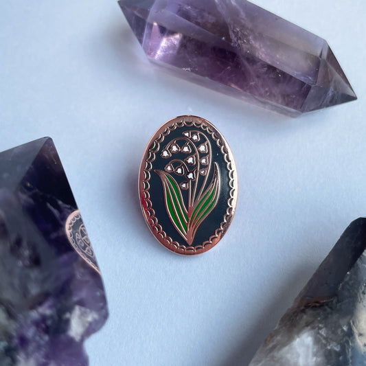 An oval shaped enamel pin with a lily of the valley flower on it surrounded by crystals.