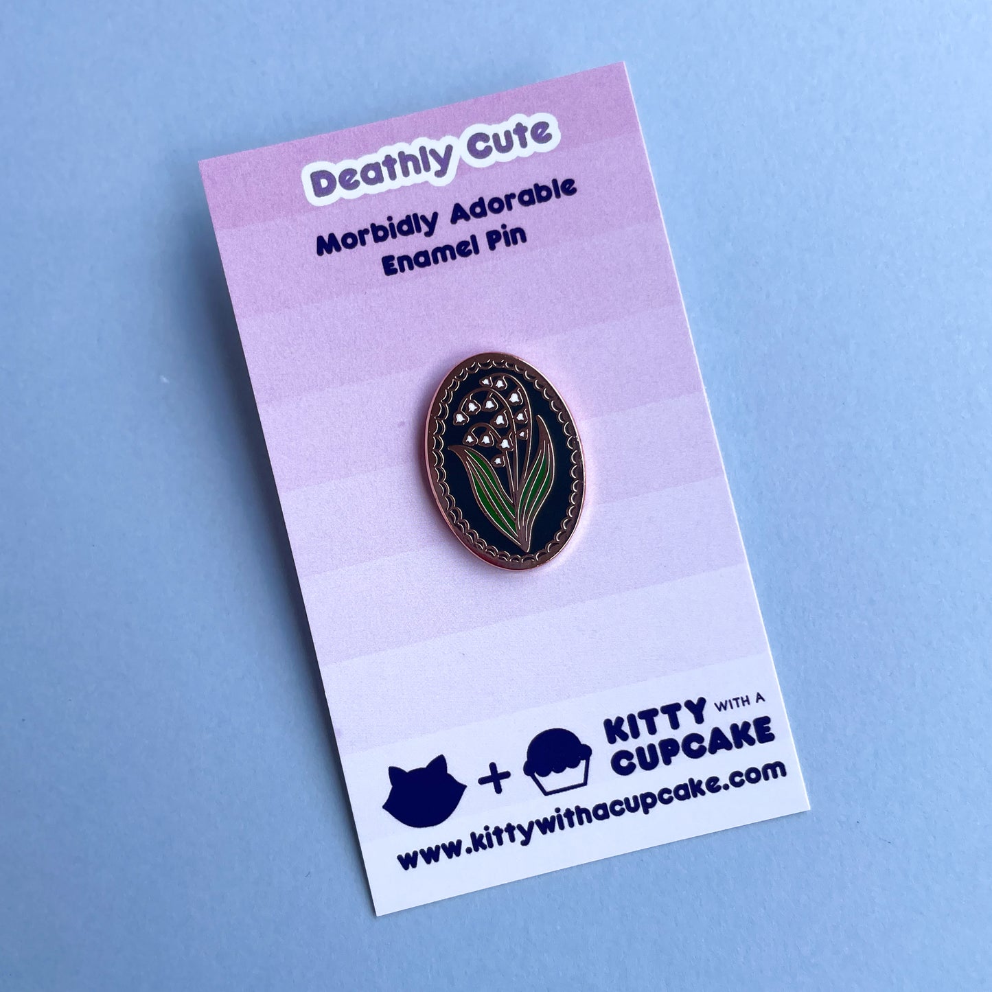An oval shaped enamel pin with a lily of the valley flower on it on a pink card. 