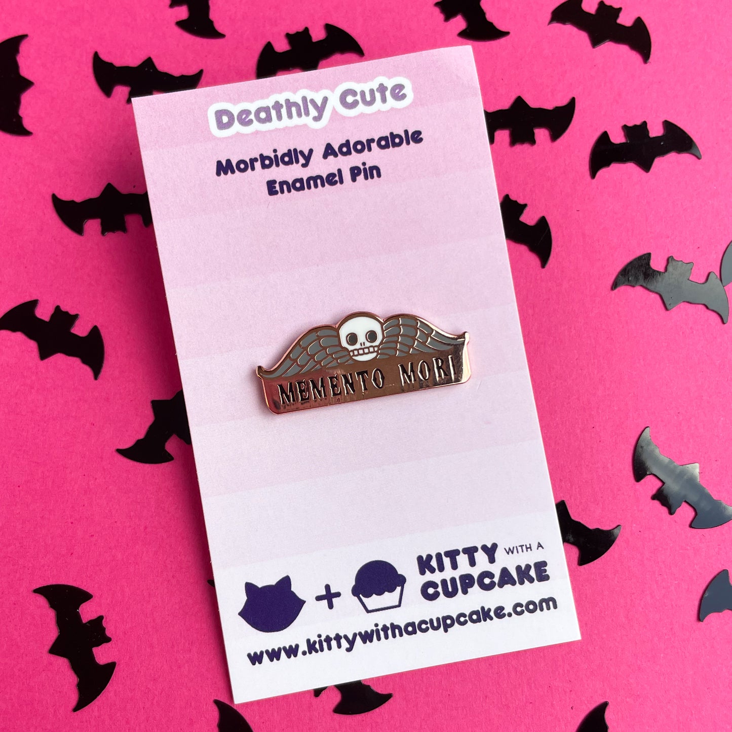 An enamel pin that reads "Memento Mori" with a skull and wings on it on a pink card. 