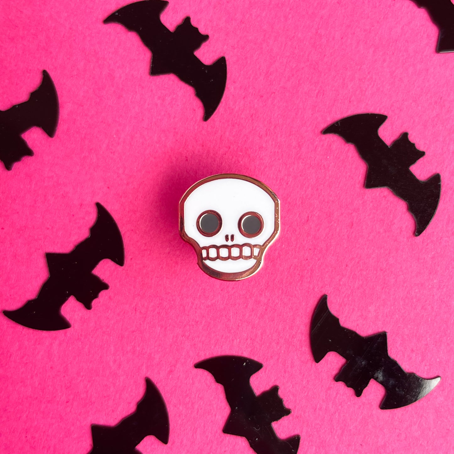 A cute skull enamel pin on a hot pink background that is covered in bat confetti. 