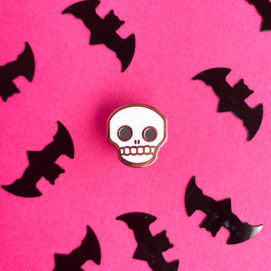 A cute skull enamel pin on a hot pink background that is covered in bat confetti.