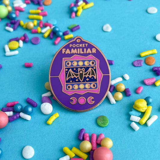 An enamel pin shaped like a Tamagotchi that reads "Pocket Familiar" at the top. It has a pink and purple shell. The screen has moon phases and a bat with night stars by it. The buttons of the tamagotchi are moon phases.  The pin is on a blue background that is covered in pastel sprinkles.