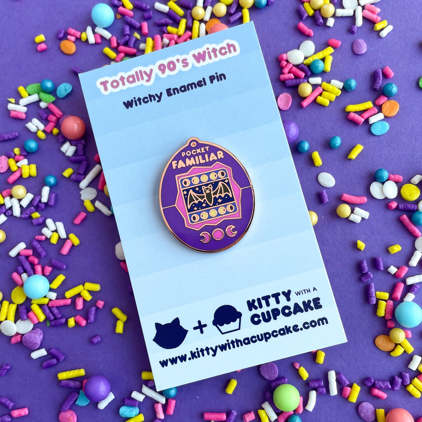 An enamel pin shaped like a Tamagotchi that reads "Pocket Familiar" at the top. It has a pink and purple shell. The screen has moon phases and a bat with night stars by it. The buttons of the tamagotchi are moon phases. The pin is on a blue backing card that reads "Totally 90's Witch" it is on a purple background covered in pastel sprinkles.