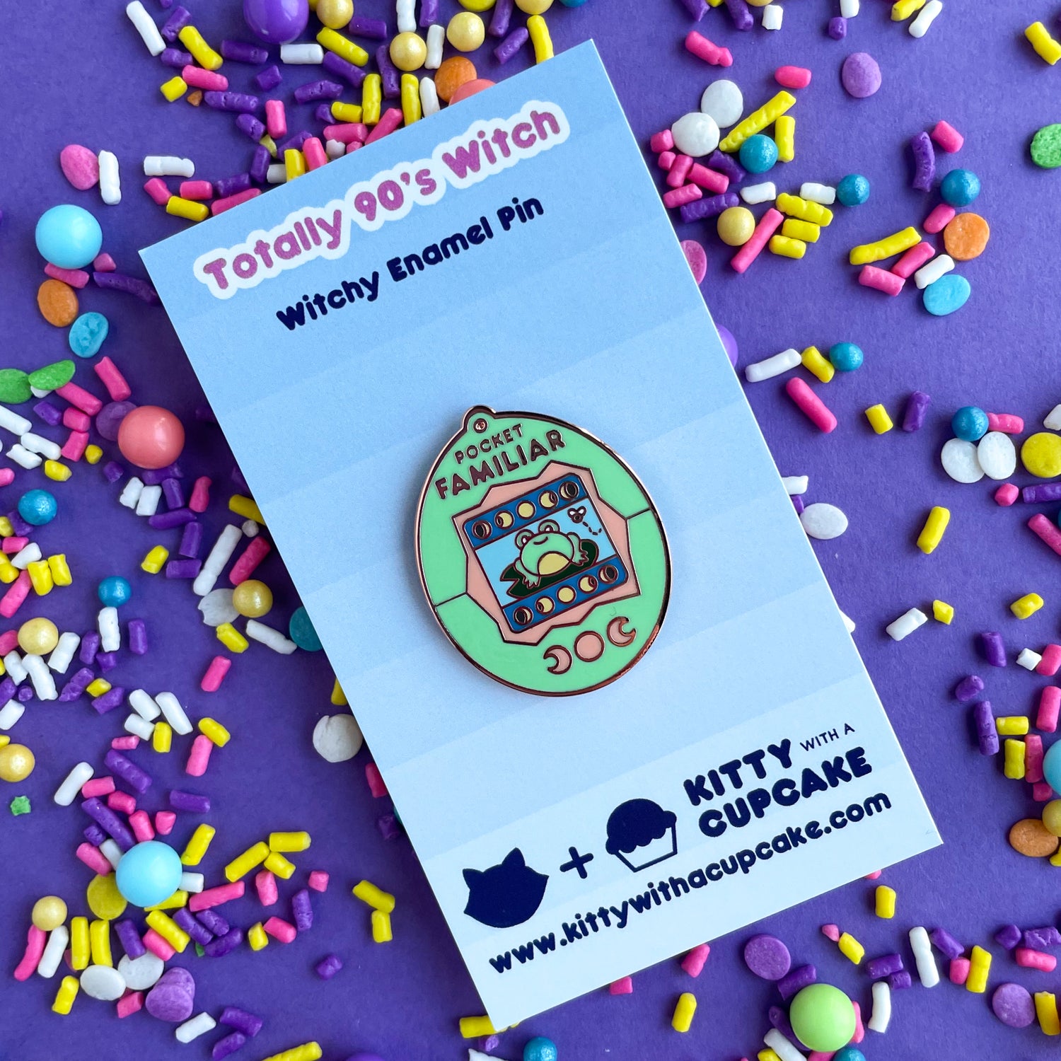 An enamel pin shaped like a Tamagotchi that reads "Pocket Familiar" at the top. It has a mint green and peach shell. The screen has moon phases and a frog on a lily pad looking at a fly. The buttons of the tamagotchi are moon phases. The pin is on a blue backing card that reads "Totally 90's Witch". This is on a purple background that is covered in pastel sprinkles.