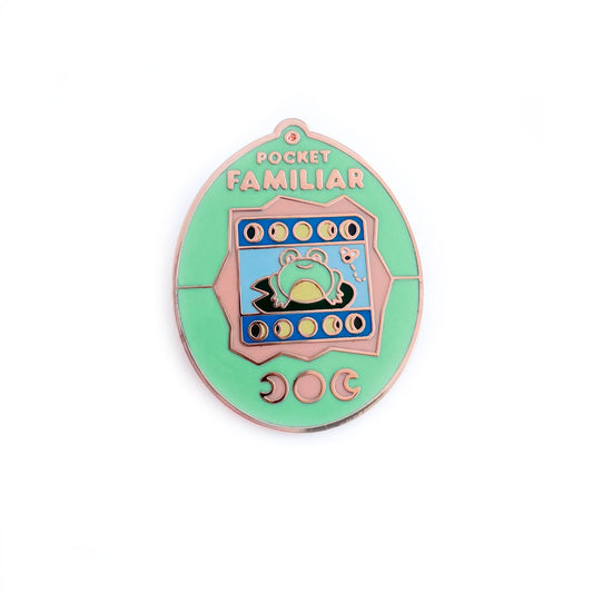 An enamel pin shaped like a Tamagotchi that reads "Pocket Familiar" at the top. It has a mint green and peach shell. The screen has moon phases and a frog on a lily pad looking at a fly. The buttons of the tamagotchi are moon phases. 