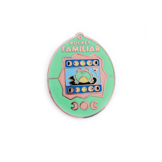 An enamel pin shaped like a Tamagotchi that reads "Pocket Familiar" at the top. It has a mint green and peach shell. The screen has moon phases and a frog on a lily pad looking at a fly. The buttons of the tamagotchi are moon phases.