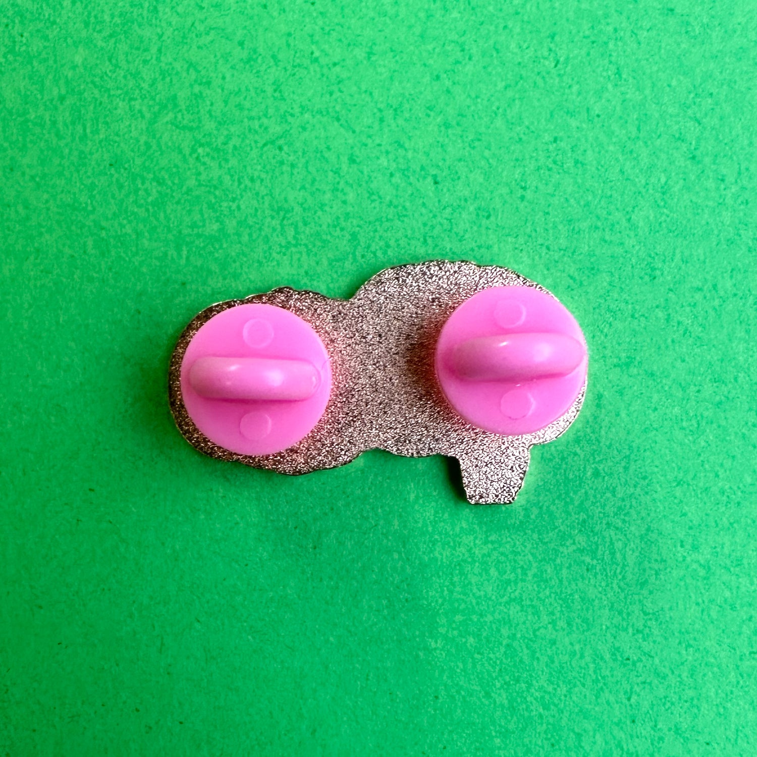The back of an enamel pin with two pink rubber pin backs on a green paper background