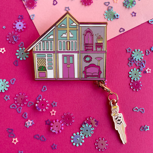 An enamel pin of a pink doll house with a Barbie shaped charm hanging from the corner on a pink paper background with confetti.