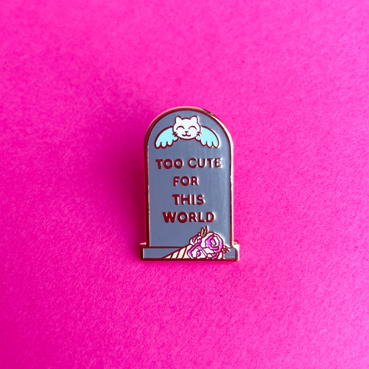 A headstone shaped enamel pin that reads "Too Cute For This World" with a cat angle on it and a bouquet by it on a pink background.