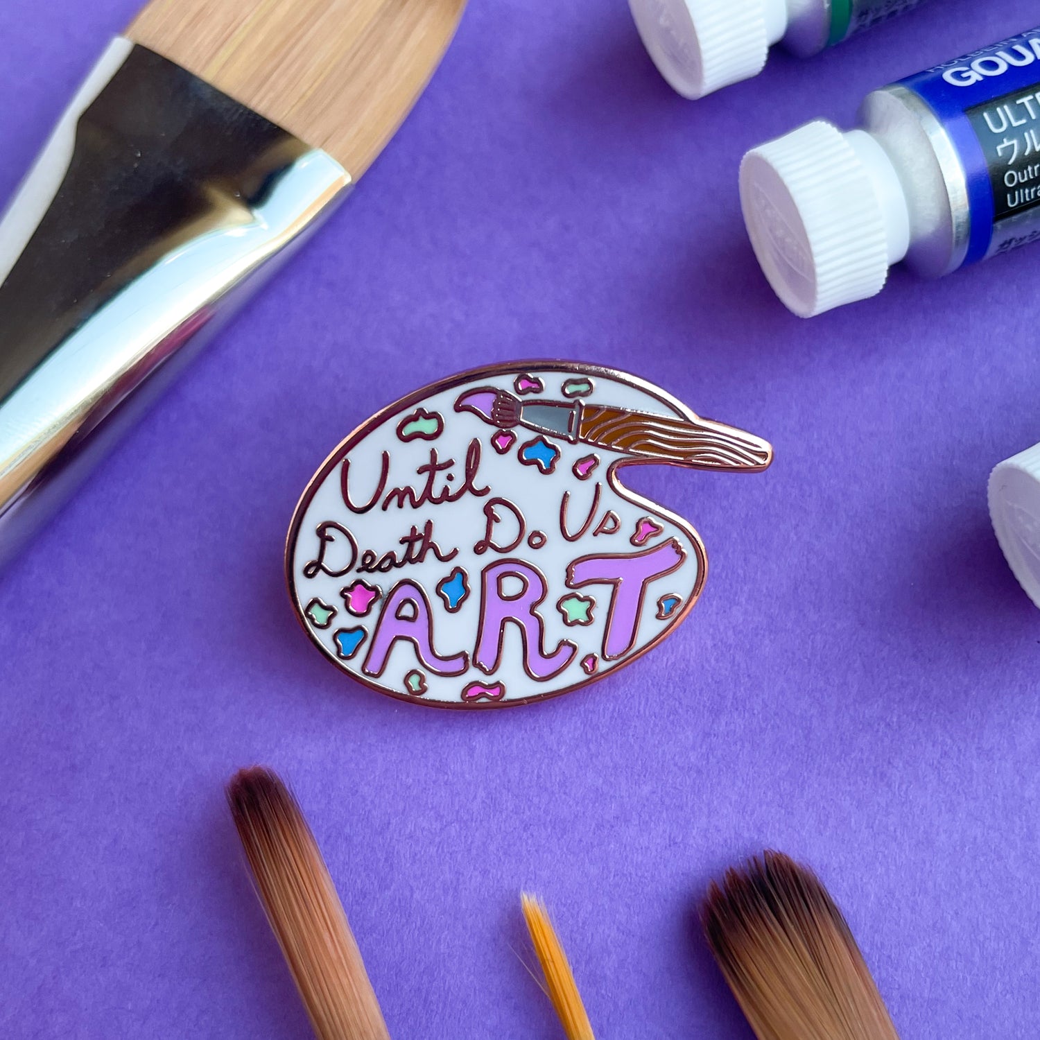 An enamel pin shaped like an artist's palette with a paint brush that reads "until death do us art" on a purple background with paintbrushes and paint around it. 