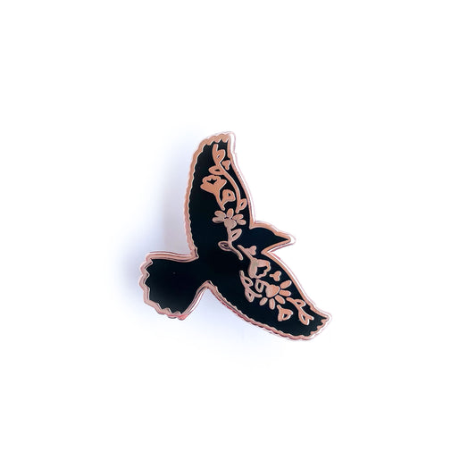 An enamel pin shaped like the silhouette of a raven with flowers along its wingspan.