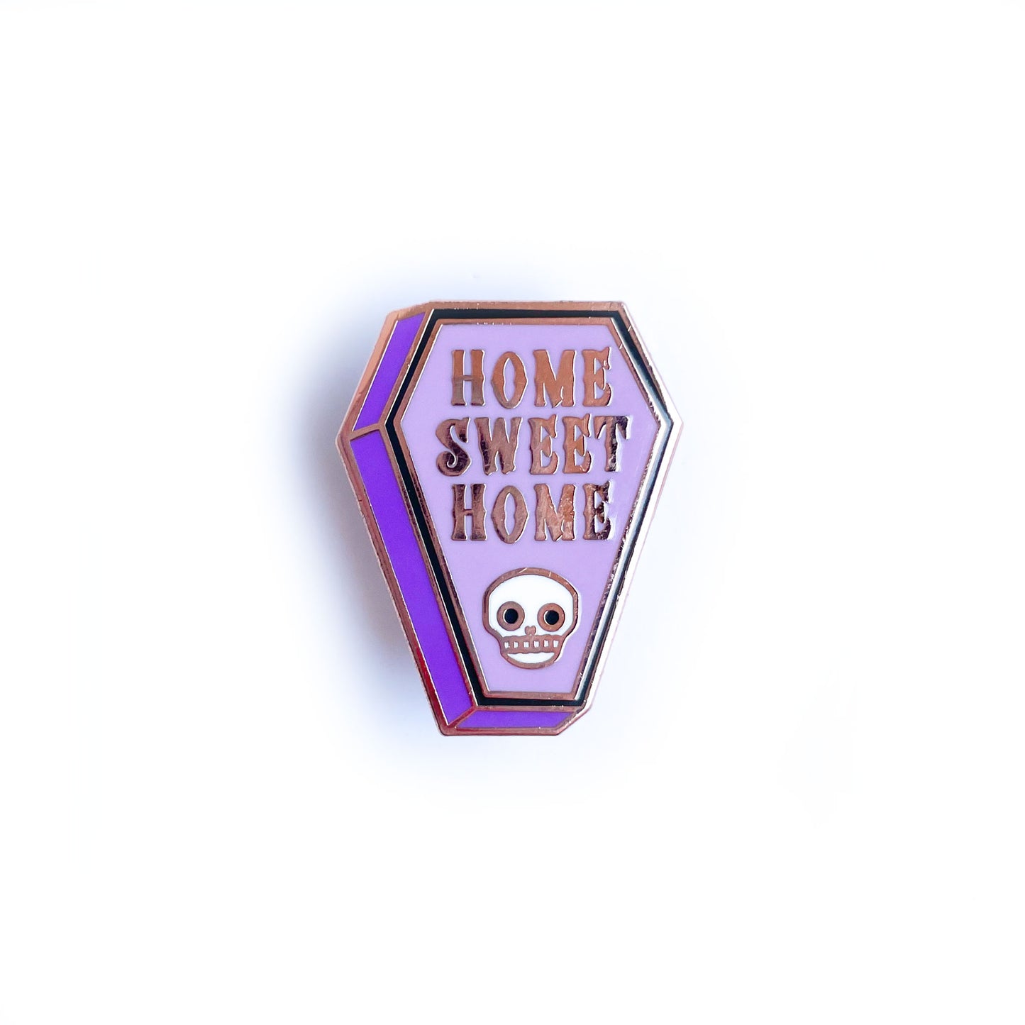 A coffin shaped enamel pin with the words "Home Sweet Home" on it and a skull.