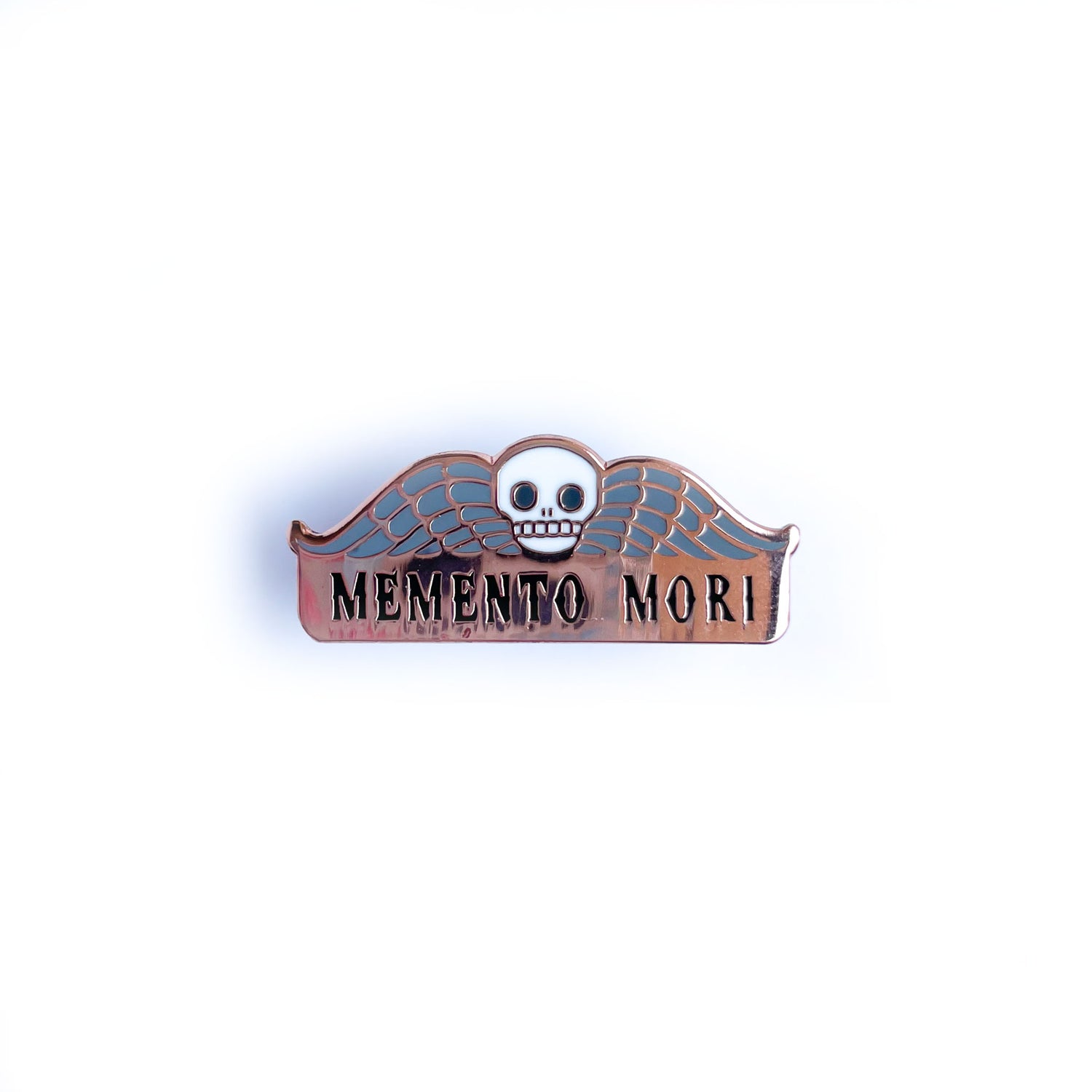 An enamel pin that reads "Memento Mori" with a skull and wings on it. 