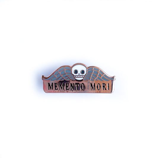 An enamel pin that reads "Memento Mori" with a skull and wings on it.