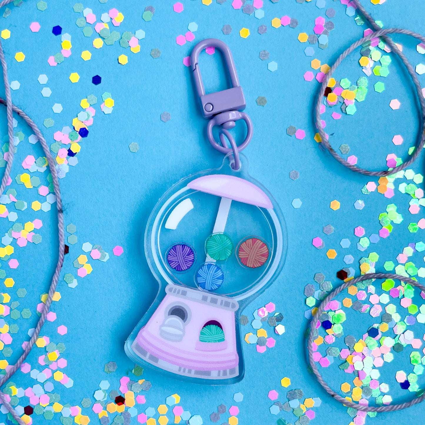 An acrylic keychain with a lavender closure that is shaped like a gumball machine that is filled with yarn balls this is on a blue background covered in yarn and confetti. 