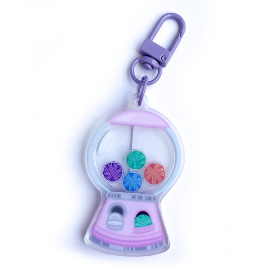 An acrylic keychain shaped like a pink gumball machine with yarn balls in it instead of gum balls. 