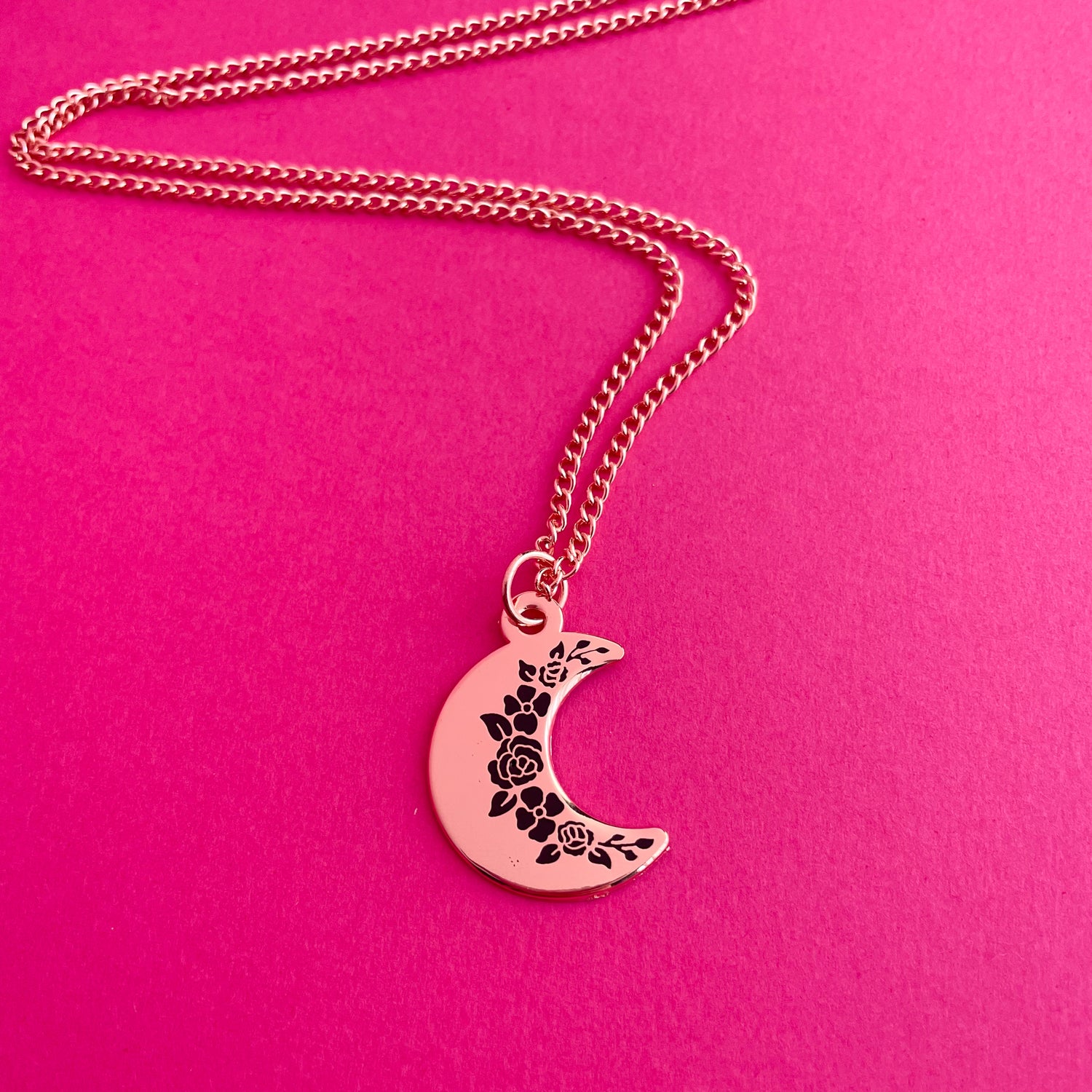 A crescent moon pendanct with black floral silhouettes along the inside edge. It has a copper colored chain. It is on a hot pink background.