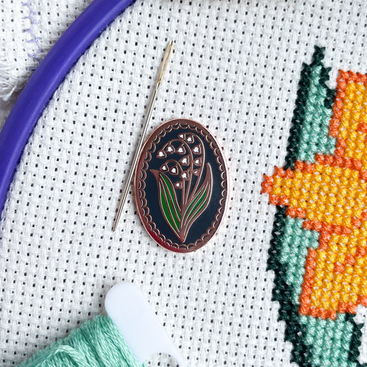 An oval needle minder with a lily of the valley on a cross stitch project 