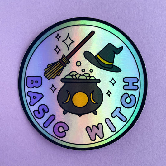 A circular holographic sticker. There is a mint green border around a pastel pink circle in the background. The bottom of the pin has bubble letters in purple that read "Basic Witch" above this there is a black cauldron with yellow moon phases full of bubbles. There is a broom above this and a black pointed hat to the right of the broom. Sparkles are around all of the objects. The sticker is on a lavender background.
