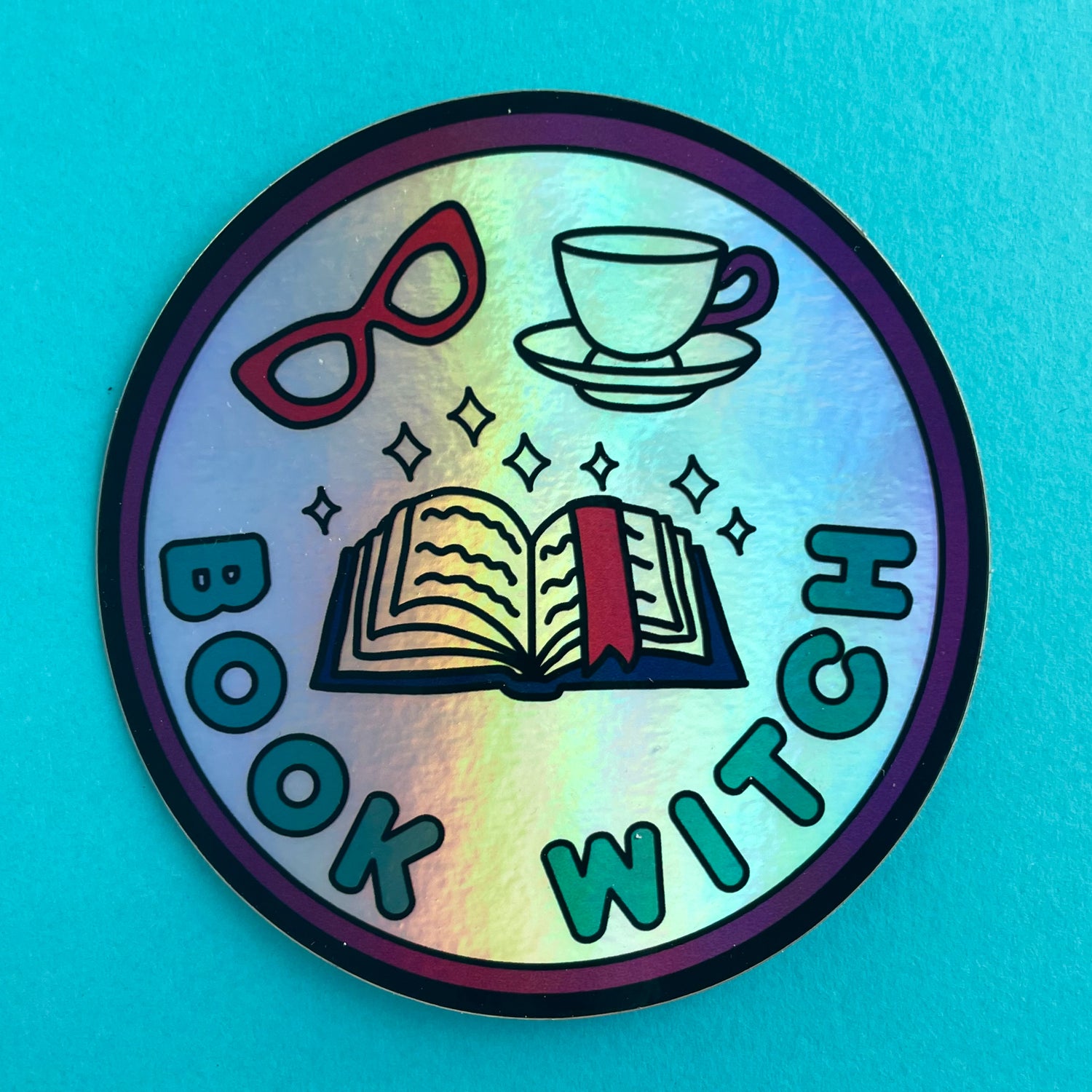 A circular holographic sticker. It has a maroon border with turquoise text that reads "Book Witch". There is an open book, sparkles, red glasses and a teacup above the words.