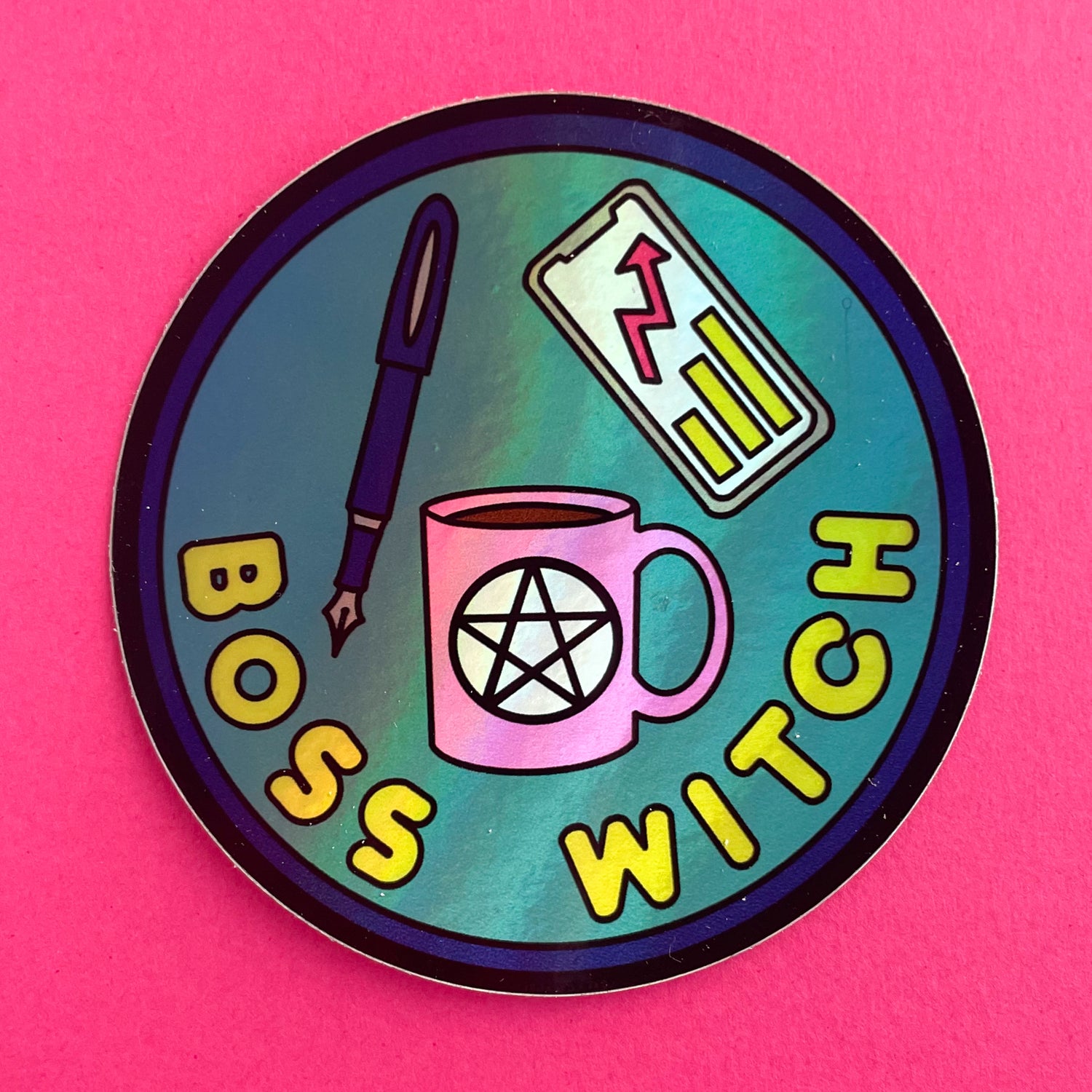 A circular holographic sticker. It has a dark purple border with lime green text that reads "Boss Witch". There is a pink coffee cup with a pentacle on it, a purple fountain pen, and a phone with a business graph depicted above the words. The sticker is on a hot pink background