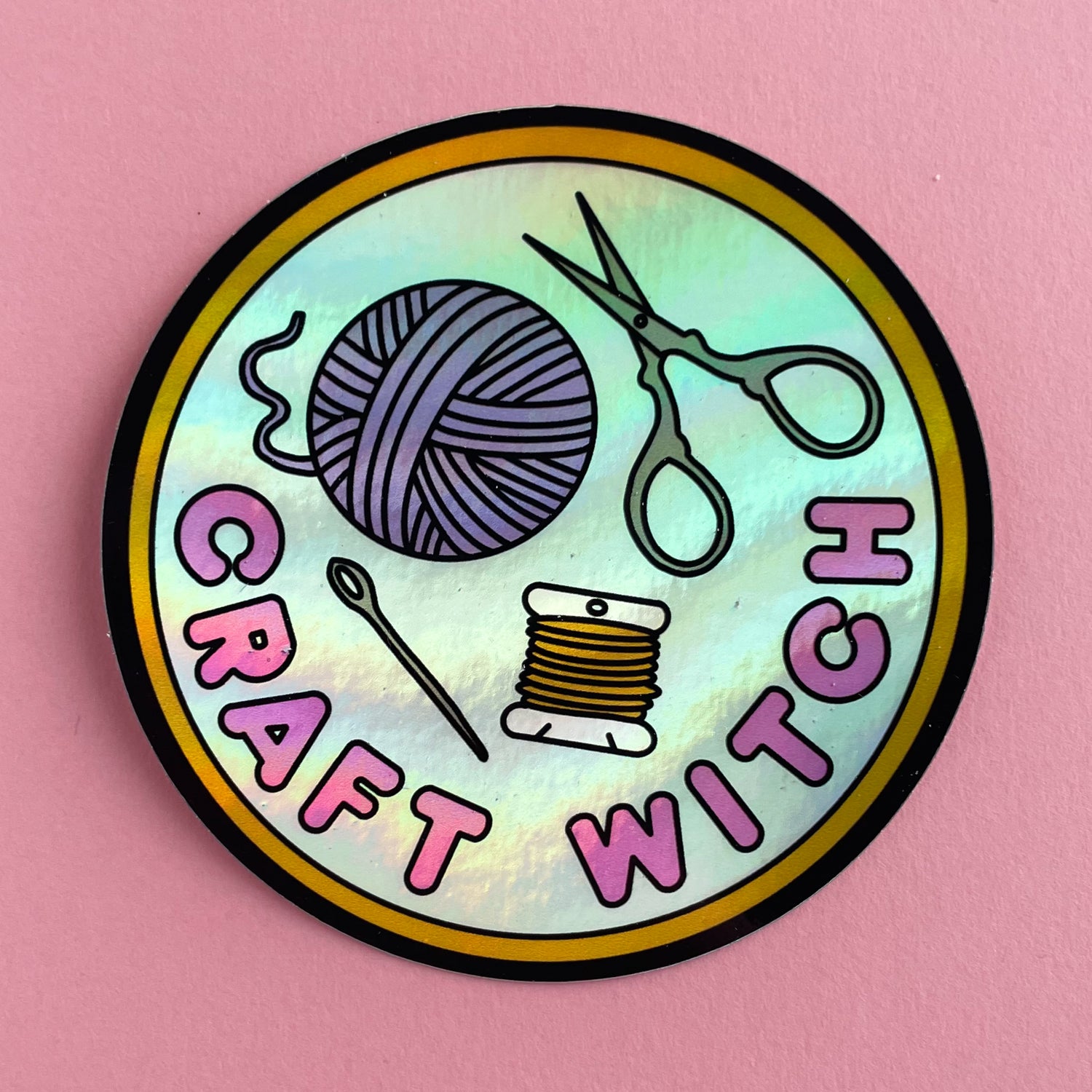 A circular holographic sticker. It has a yellow borer with a light blue background and hot pink text that reads "Craft Witch". Above the words a needle, thread, yarn ball, and scissors are depicted.