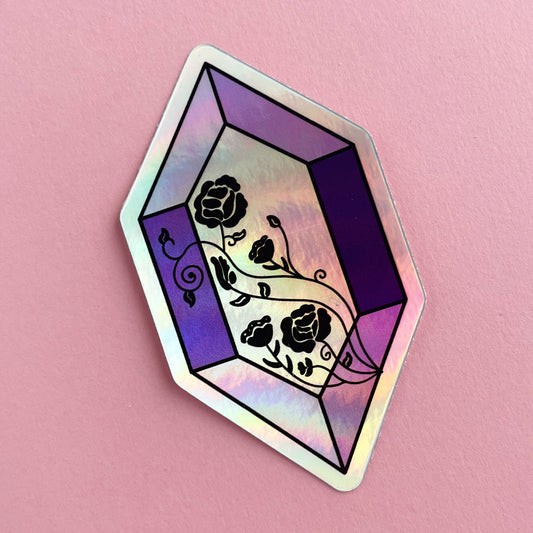 A holographic sticker in the shape of an elongated rectangle. It is various shades of purple and has roses in black line art across the face. The sticker is on a pink background.