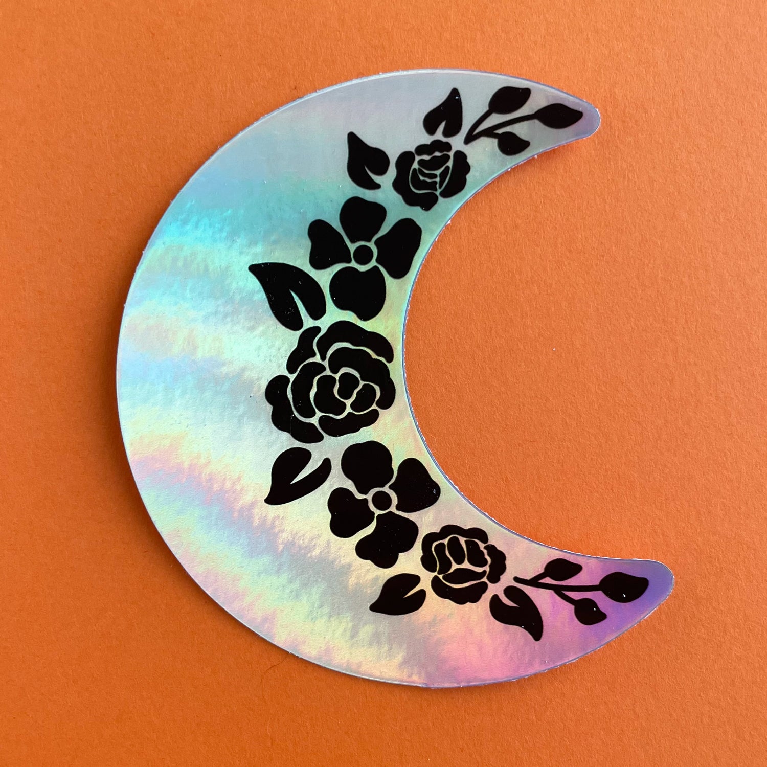 A crescent moon shaped holographic sticker. It has black floral outlines along the inside edge. The sticker is on an orange background.