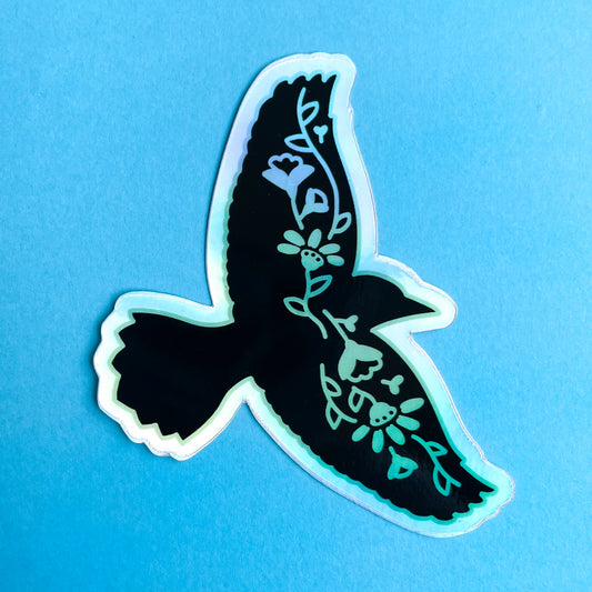 A holographic sticker shaped like the silhouette of a raven with flowers along its wingspan. The sticker is on a blue background