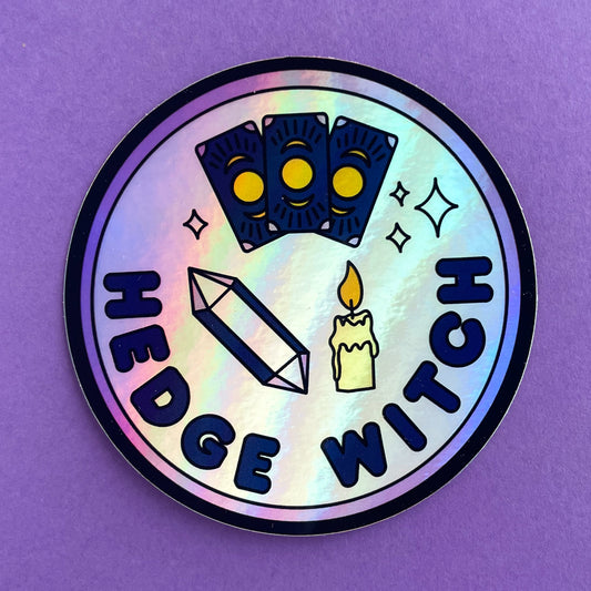 A circular holographic sticker with a purple border and words that read "Hedge Witch". It has an amethyst crystal, a candle, and tarot cards depicted on it. The sticker is on a purple background.