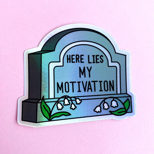 A gravestone shaped sticker that reads "Here Lies My Motivation" with lily of the valley flowers in front of it on a light pink background