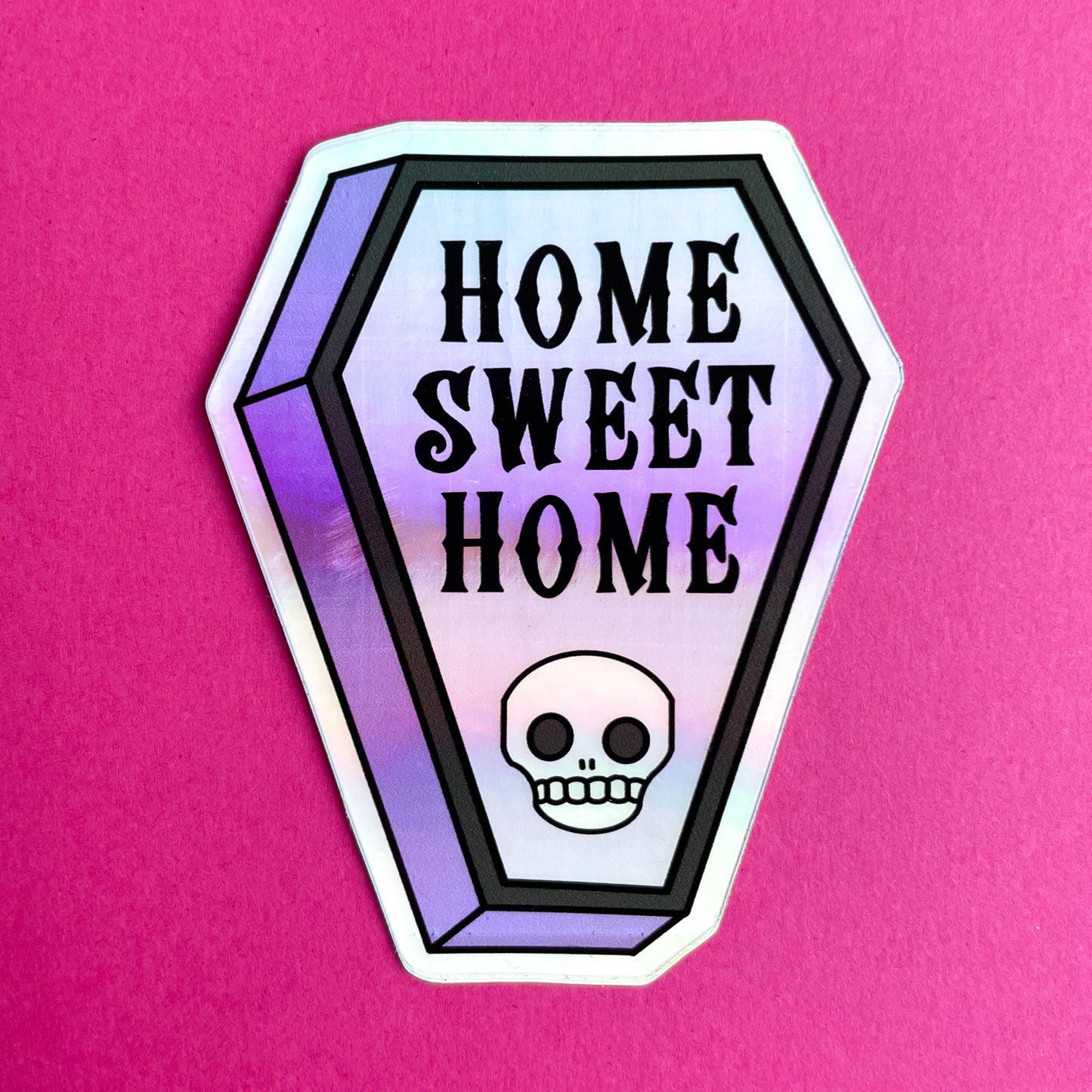 A coffin shaped holographic sticker that reads "Home Sweet Home" and has a skull on it on a hot pink background. 