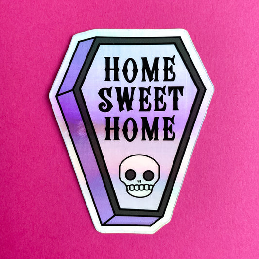 A coffin shaped holographic sticker that reads "Home Sweet Home" and has a skull on it on a hot pink background.
