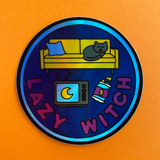 A circular holographic sticker with a blue border, navy background, and maroon words that read "Lazy Witch". It has a vintage TV, bag of chips, and a yellow couch with a black cat on it. The sticker is on a orange background.
