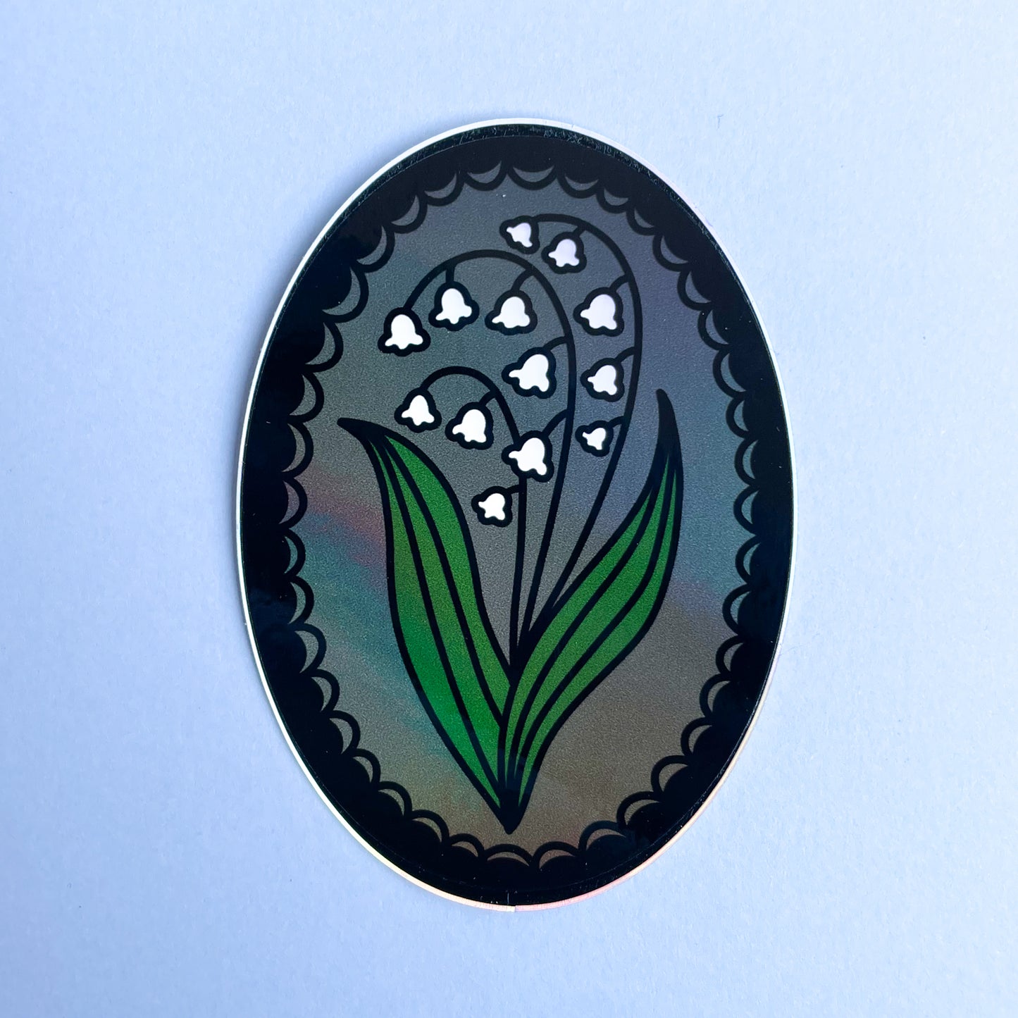 An oval shaped sticker with a lily of the valley in a lace frame on a grey background.
