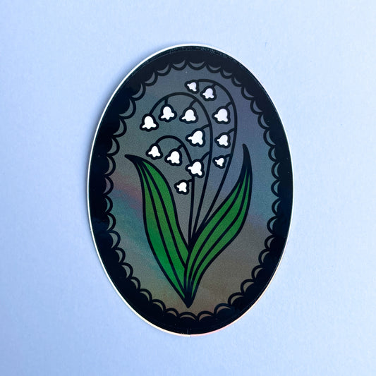 An oval shaped sticker with a lily of the valley in a lace frame on a grey background.