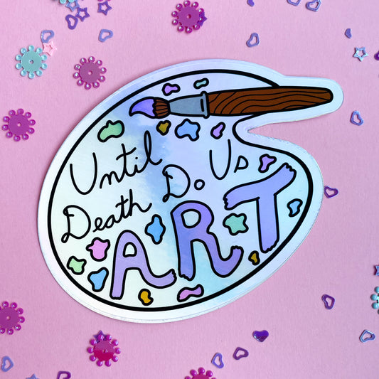 A holographic sticker shaped like an artist's palette with a paint brush that reads "until death do us art". The sticker is displayed on a pink background