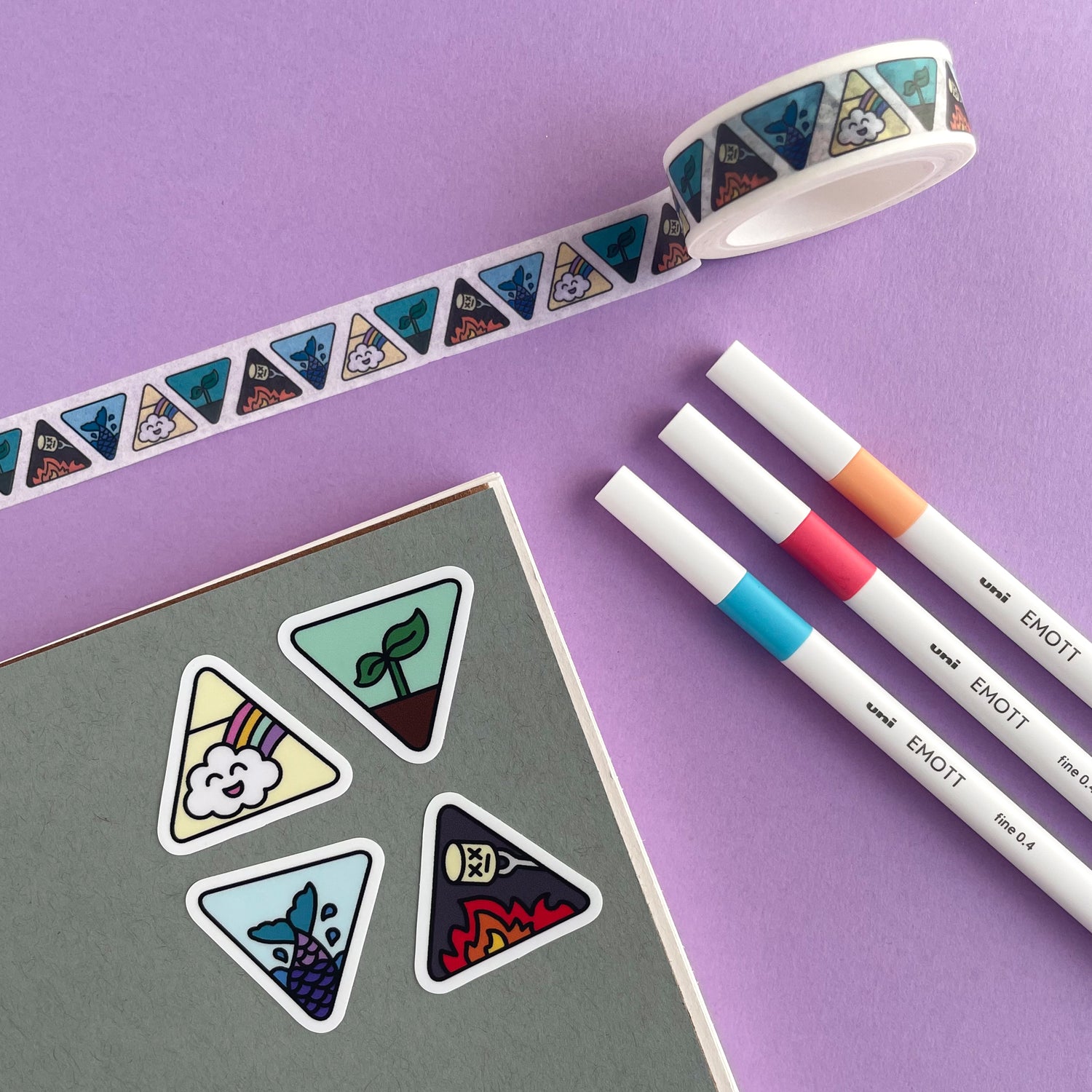 A grey notebook with stickers on it that are triangle shaped and have different cute representations of the four elements on them. There are some white pens and a roll of washi tape that matches the stickers around the notebook. All of this is on a lavender background.