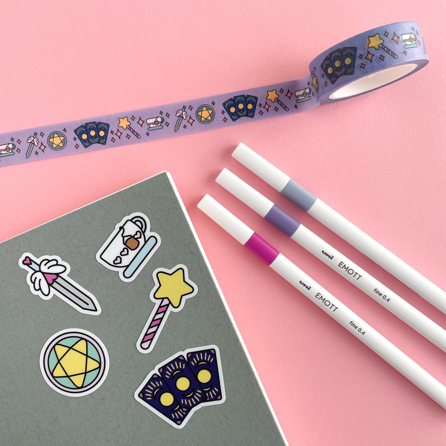 A grey notebook cover with stickers that are cute shapes of the different tarot suits, pentacles, swords, cups, wands, and fanned out cards. There are white pens and a roll of washi tape that matches the stickers around the notebook. This is all on a pink background.