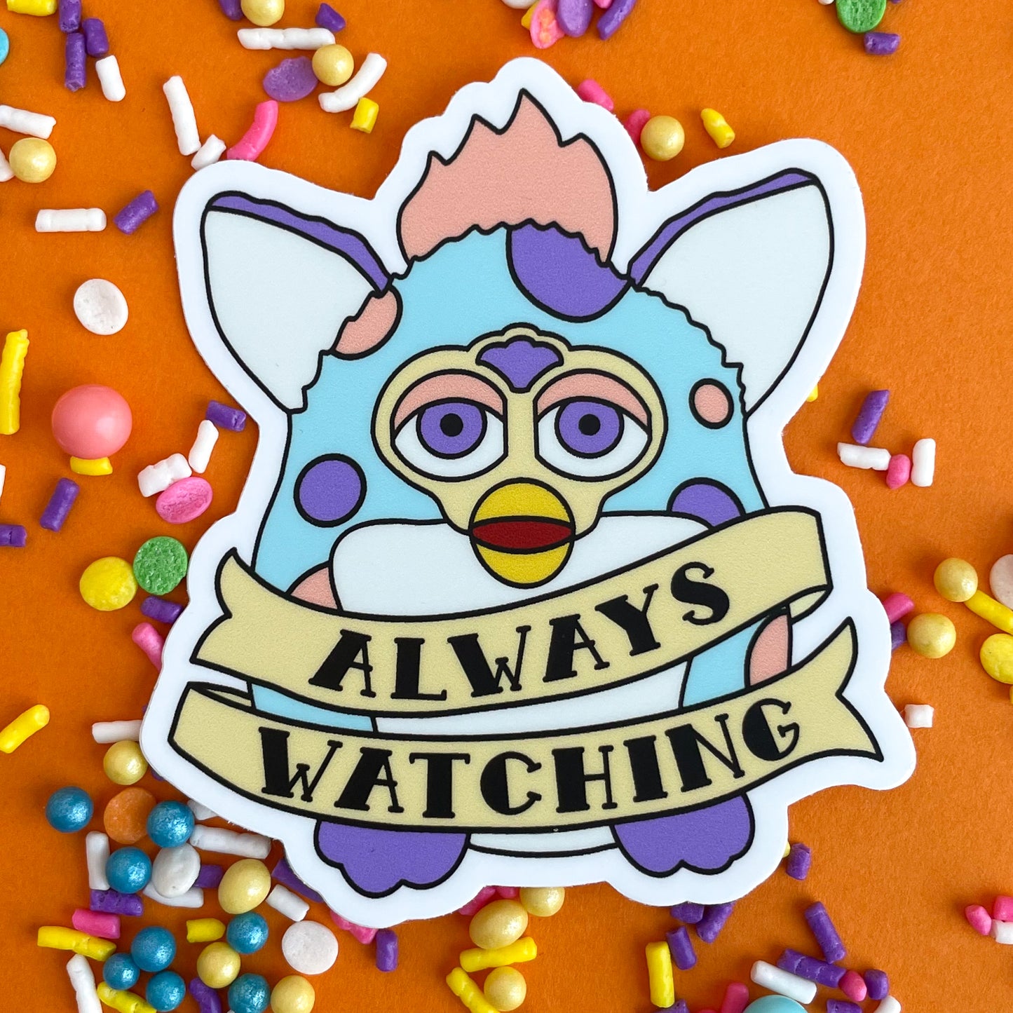 A sticker of a furby that is pastel blue with peach and purple spots. There is a tattoo style banner around him that says. "Always Watching". The sticker is on an orange background that is covered in pastel sprinkles.