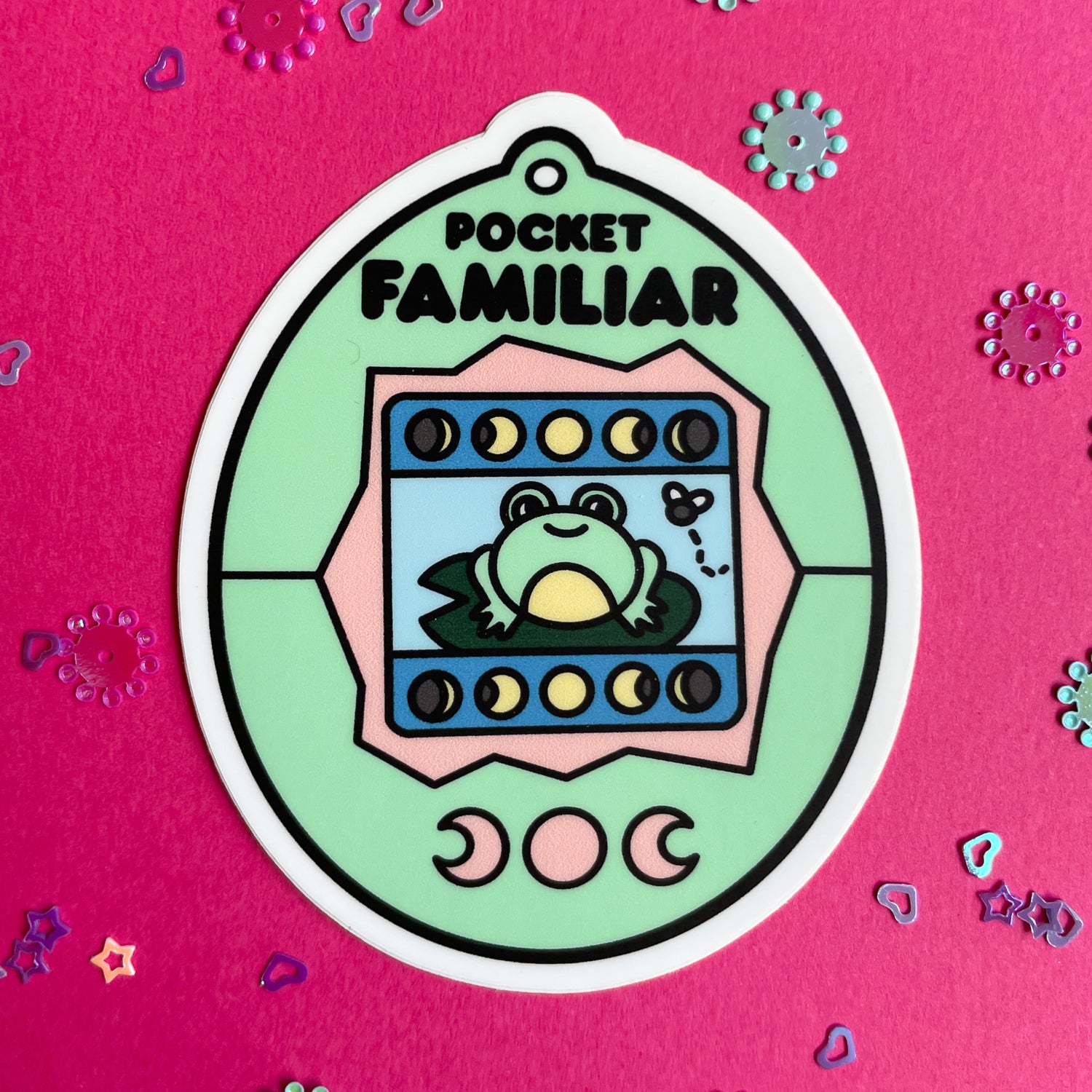 A vinyl sticker shaped like a virtual pet with a frog on the screen that has a mint and peach colored shell. The sticker is on a hot pink paper background.