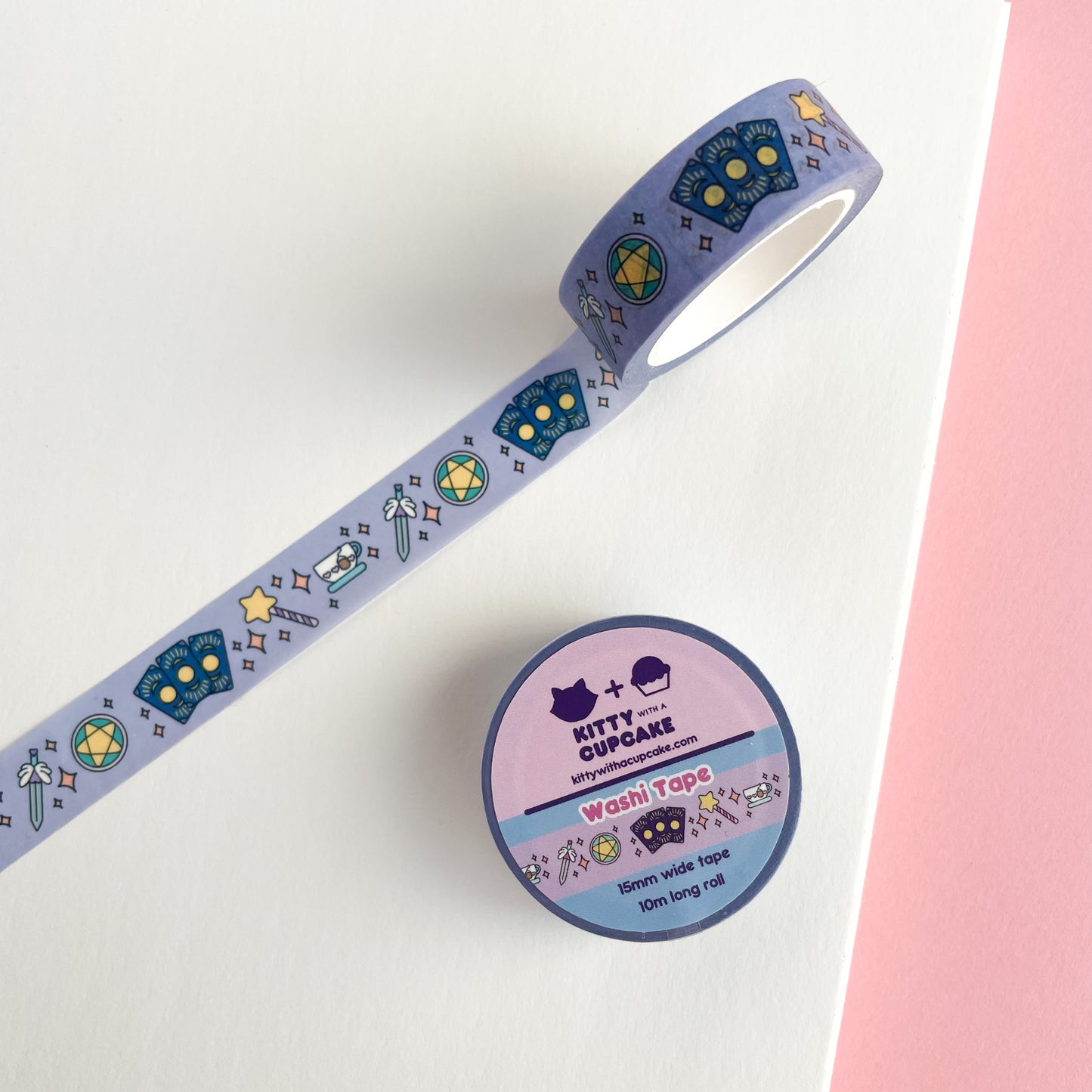 A washi tape roll. The tape features small tarot cards, a star wand, a teacup, a sword, and a pentacle all in pastel colors. There are orange pastel sparkles in between the items on a lavender background.