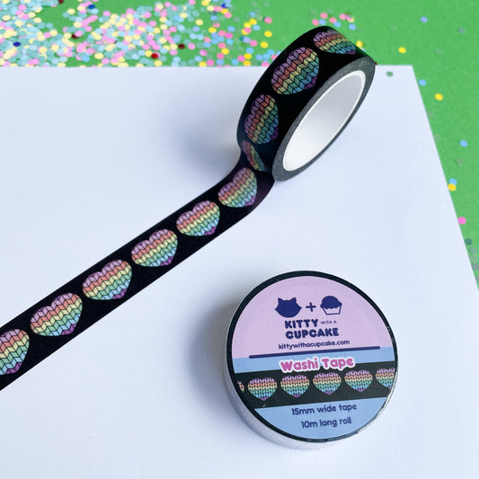 A roll of washi tape stuck to a piece of paper. The tape has a black background with pastel rainbow hearts on it. 