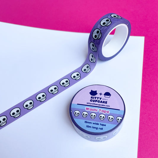 A circular roll of washi tape next to a roll of washi tape stuck to a piece of paper. The tape has a periwinkle background with skulls on it.