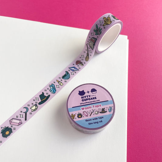 A roll of washi tape with a pink background with a collection of witchy items on it like a crystal cluster, cauldron, sickle, teacup, witch hat, and a candle. The tape roll is on a piece of paper on a hot pink background.