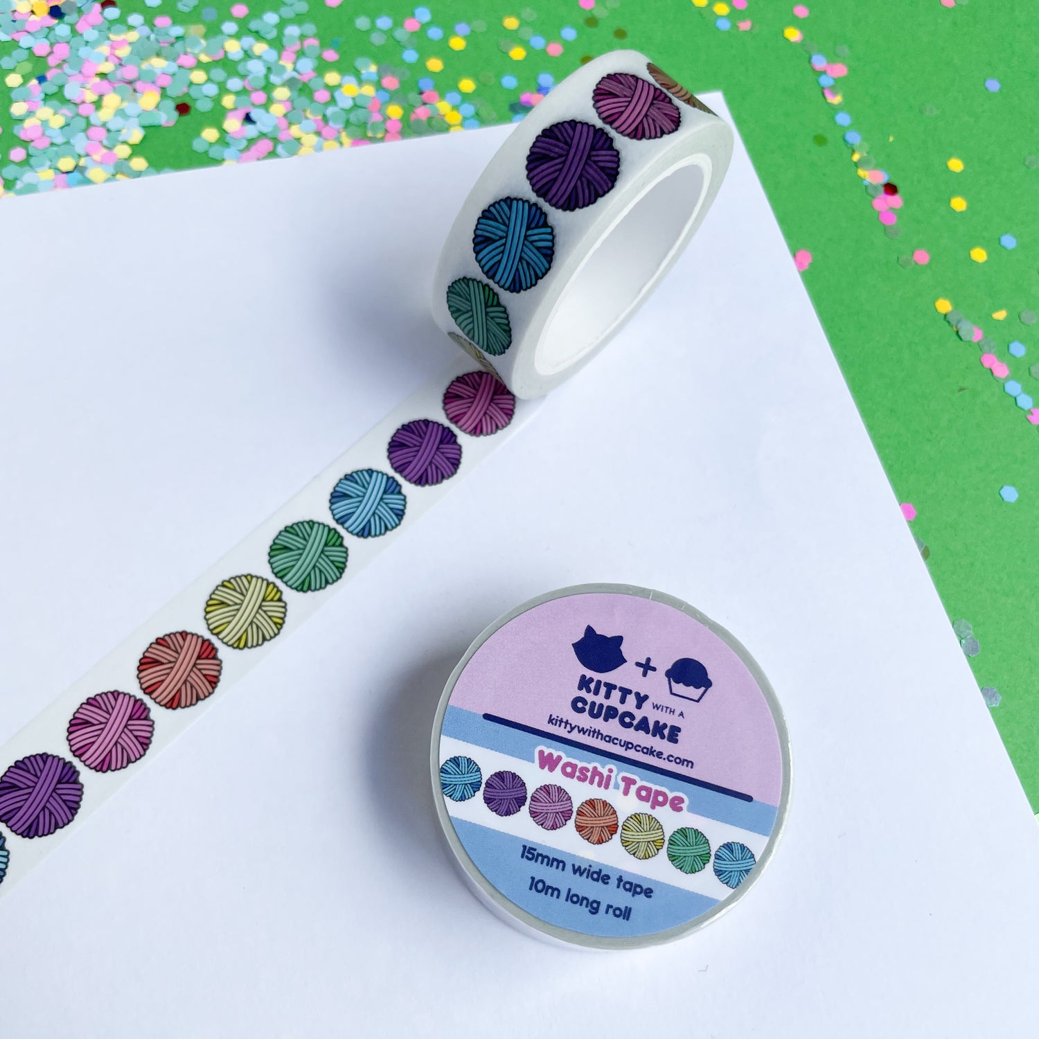 A washi tape roll on a piece of paper. The tape has a white background with pastel rainbow yarn balls. This is on a green paper background with confetti. 